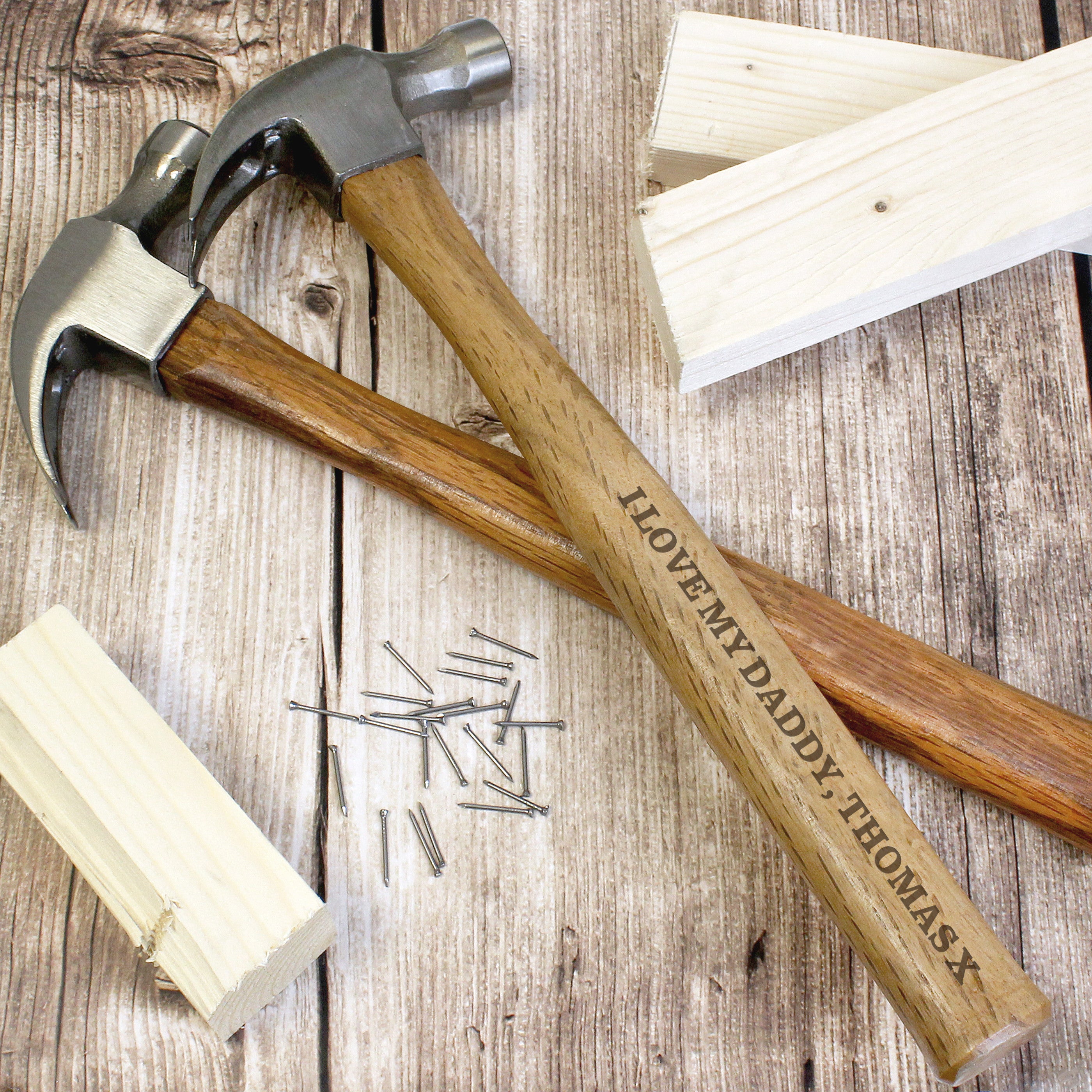 A personalised hammer with a wooden handle and metal head, engraved with the text 'Daddy', along with a few nails and pieces of wood scattered around it.