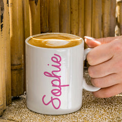 A white ceramic mug with the name 'Sophie' printed in pink script font on the side, held by a hand against a bamboo-themed backdrop.