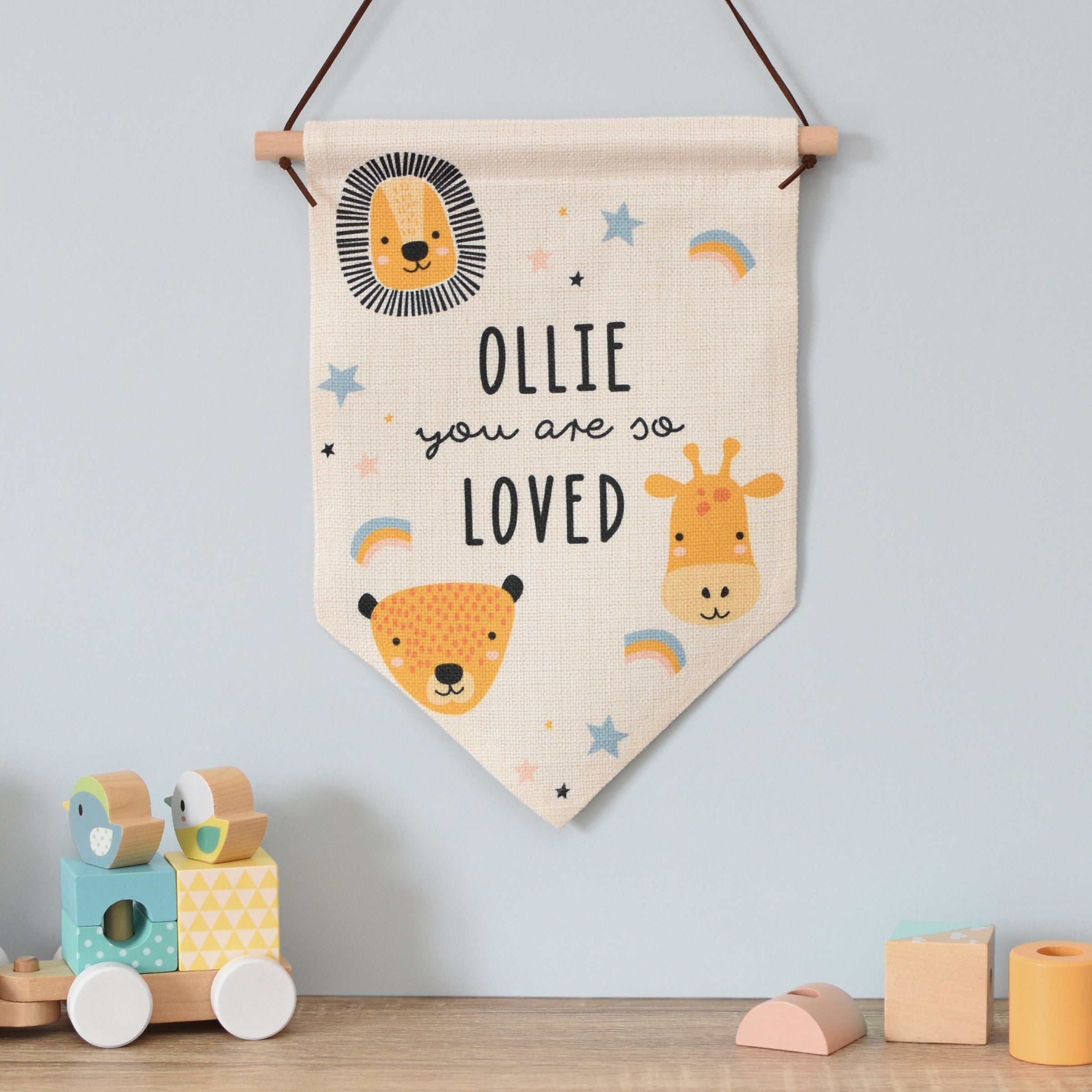 A beige-coloured hanging banner with a safari animal theme, including a lion, giraffe, and monkey, with the text 'OLIE you are so loved' displayed. 01