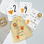 A set of personalised milestone cards with safari animal illustrations, a drawstring bag, and a small wooden clip, all laid out on a grey fabric background.
