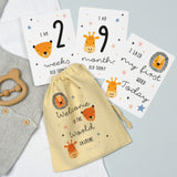 A set of personalised milestone cards with safari animal illustrations, a drawstring bag, and a small wooden clip, all laid out on a grey fabric background.
