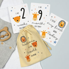 A set of personalised milestone cards with safari animal illustrations, a drawstring bag, and a small wooden clip, all laid out on a grey fabric background.