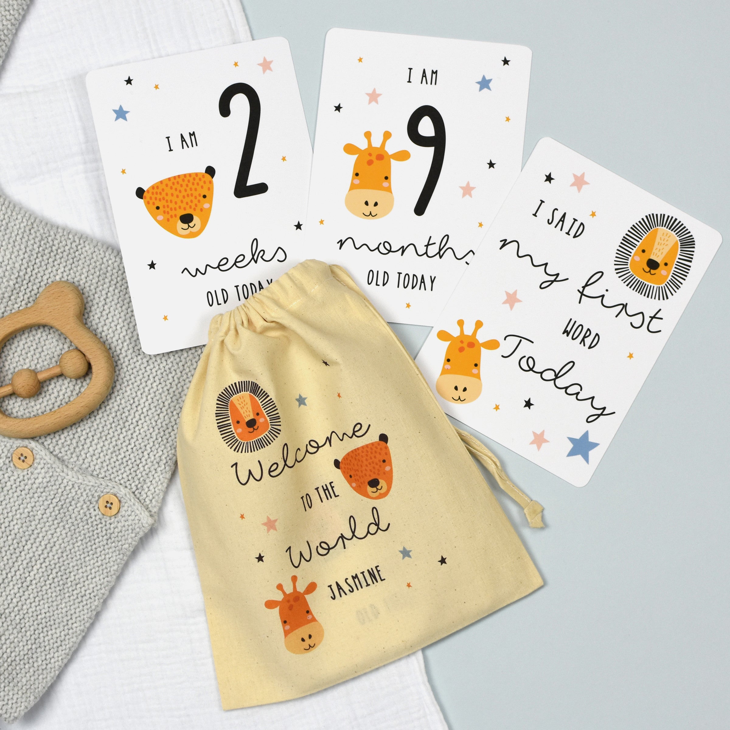 A set of personalised milestone cards with safari animal illustrations, a drawstring bag, and a small wooden clip, all laid out on a grey fabric background.