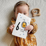 A set of personalised milestone cards with safari animal illustrations, a drawstring bag, and a small wooden clip, all laid out on a grey fabric background.