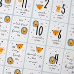 A set of personalised milestone cards with safari animal illustrations, a drawstring bag, and a small wooden clip, all laid out on a grey fabric background.
