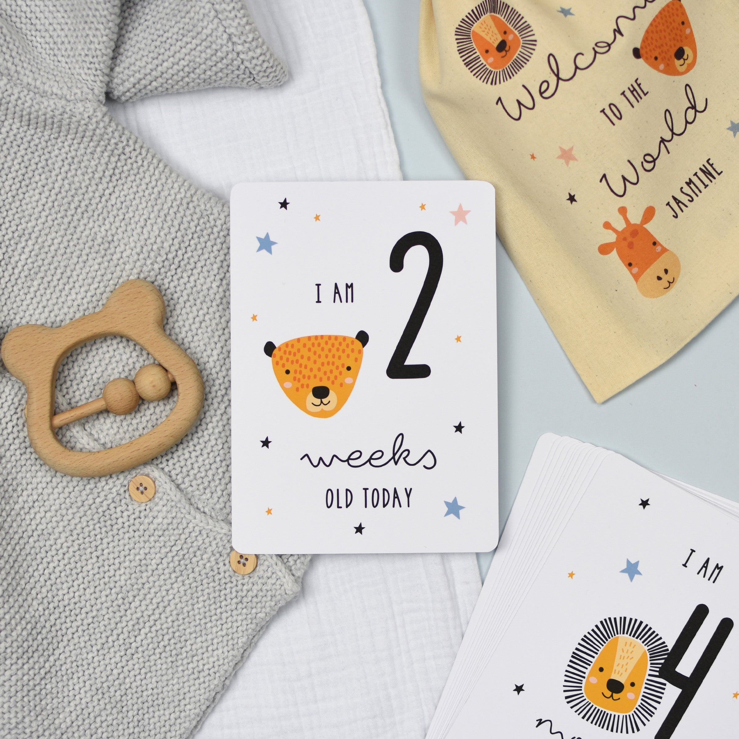 A set of personalised milestone cards with safari animal illustrations, a drawstring bag, and a small wooden clip, all laid out on a grey fabric background.