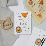A set of personalised milestone cards with safari animal illustrations, a drawstring bag, and a small wooden clip, all laid out on a grey fabric background.