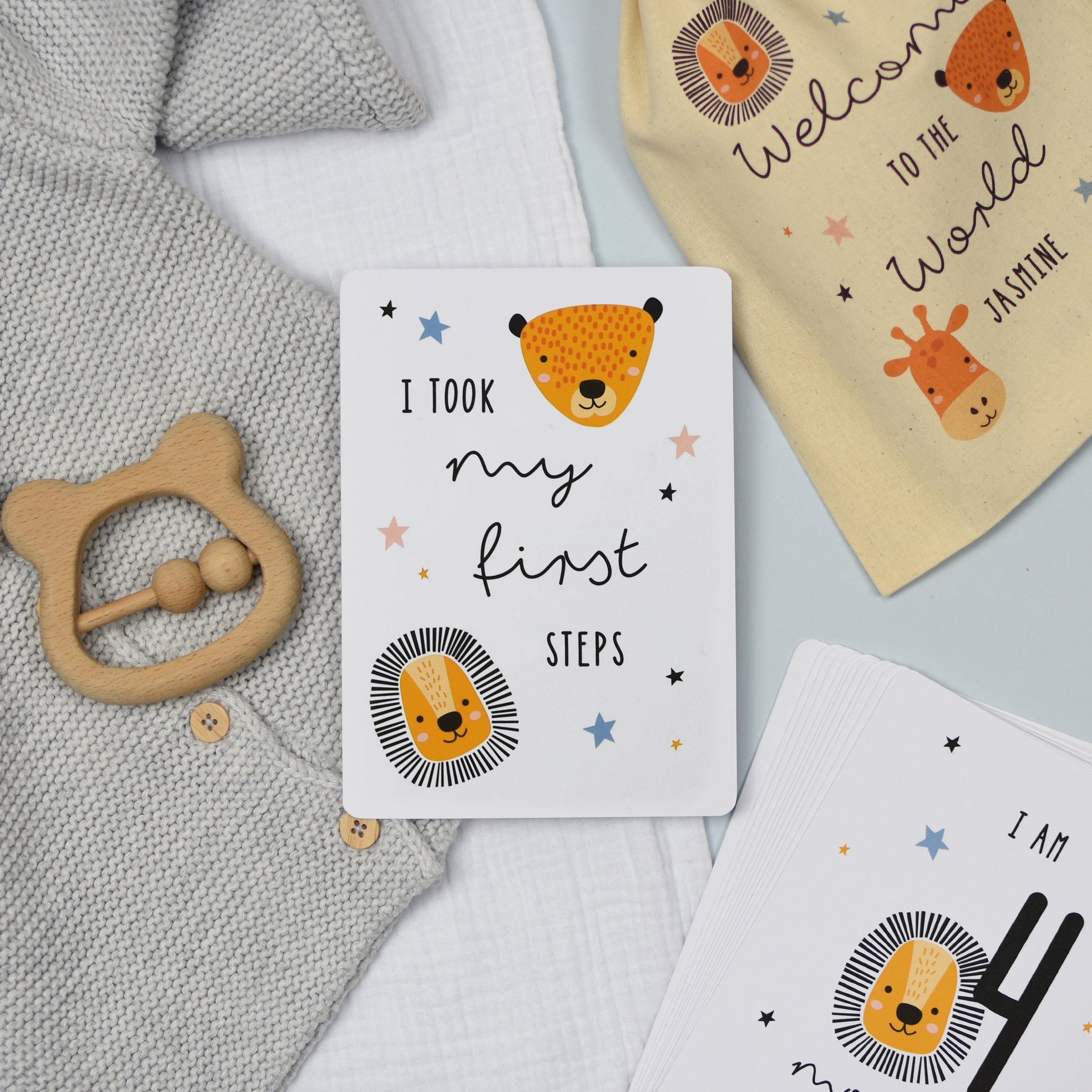 A set of personalised milestone cards with safari animal illustrations, a drawstring bag, and a small wooden clip, all laid out on a grey fabric background.