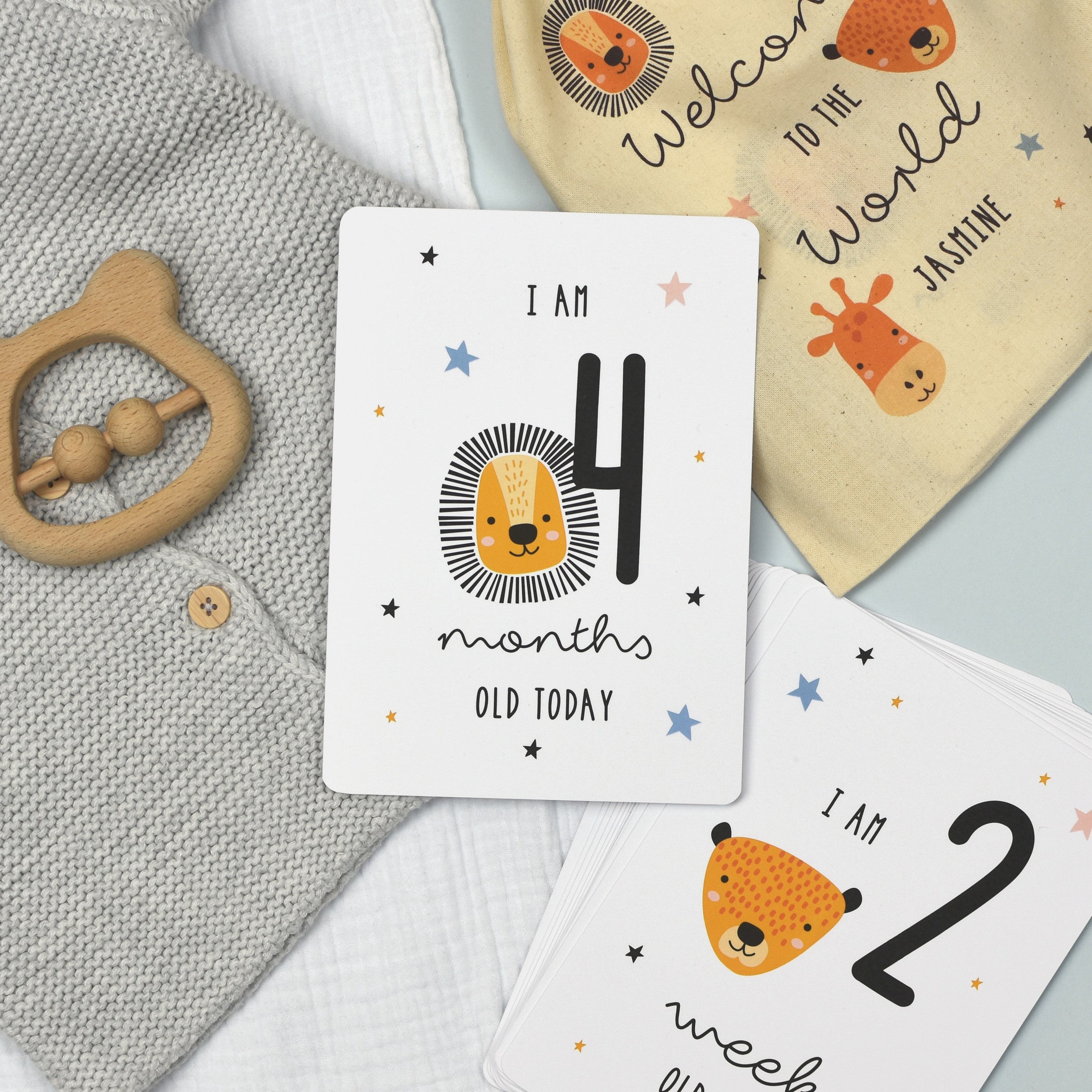 A set of personalised milestone cards with safari animal illustrations, a drawstring bag, and a small wooden clip, all laid out on a grey fabric background.