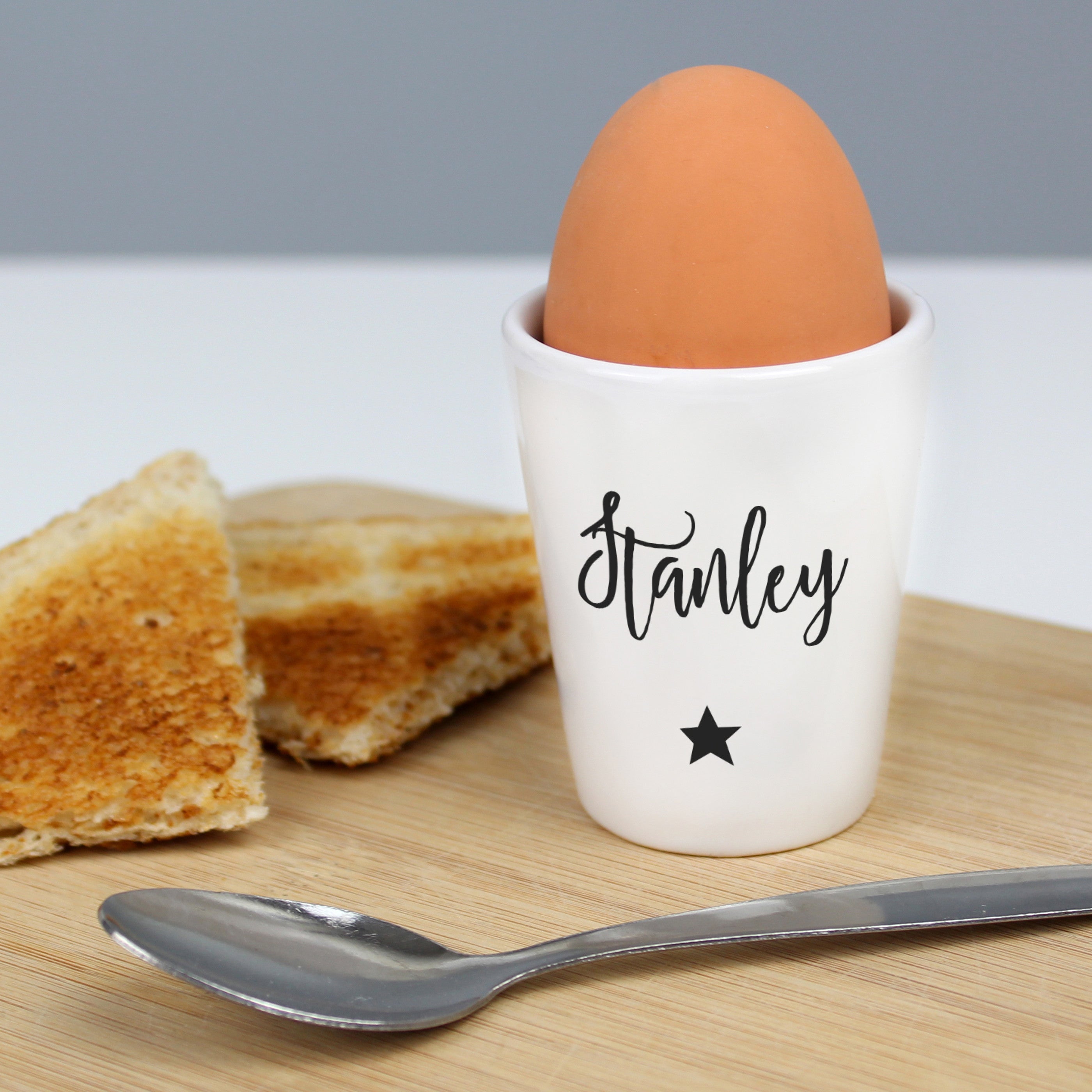 A white ceramic egg cup with the name 'Stanley' printed on it, alongside a spoon and a piece of toast, with an egg placed in the egg cup.