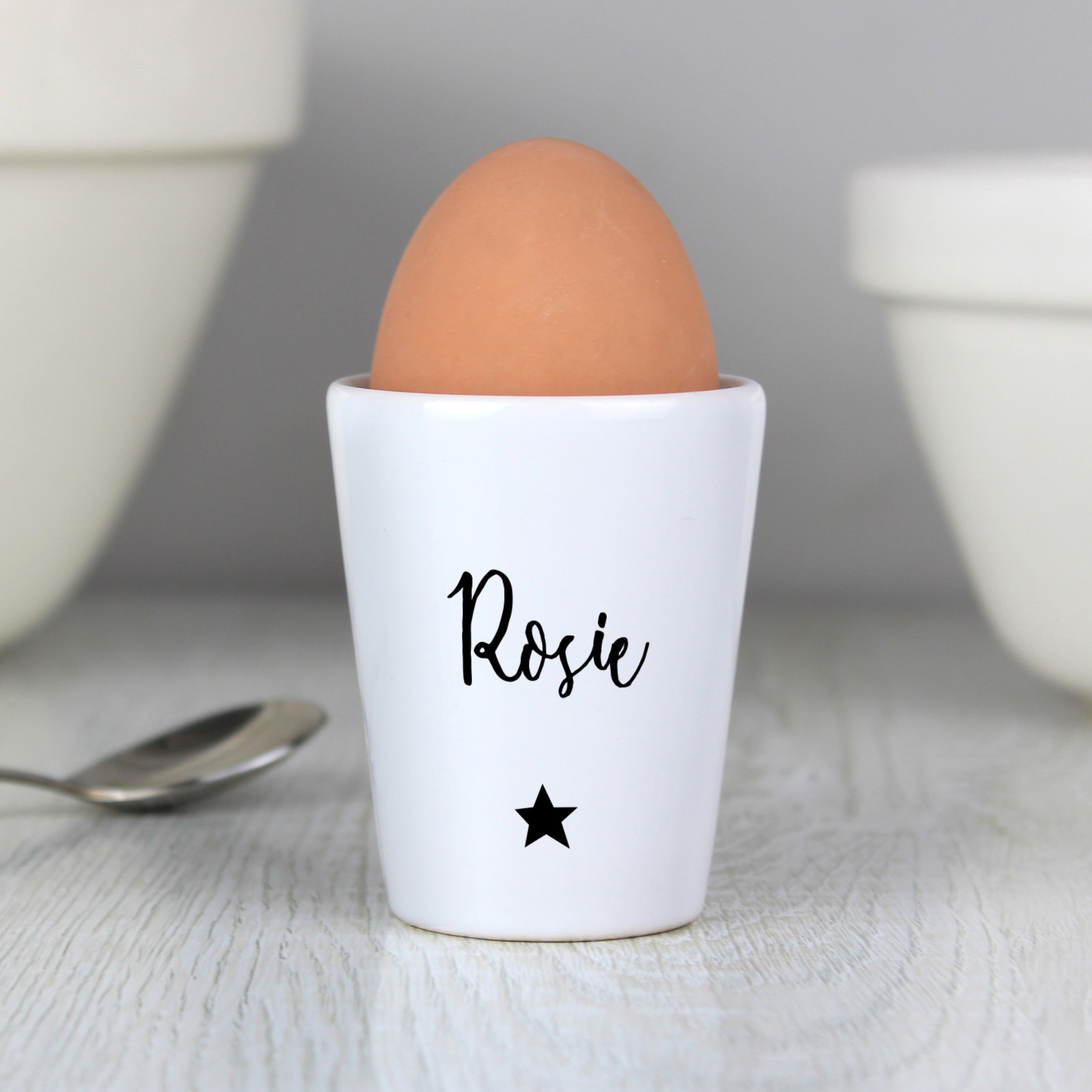 A white ceramic egg cup with the name 'Stanley' printed on it, alongside a spoon and a piece of toast, with an egg placed in the egg cup.