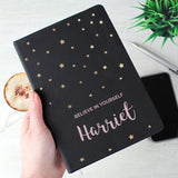 A personalised black hardback notebook with star patterns on the cover, held in a hand, with a mobile phone and a plant in the background.