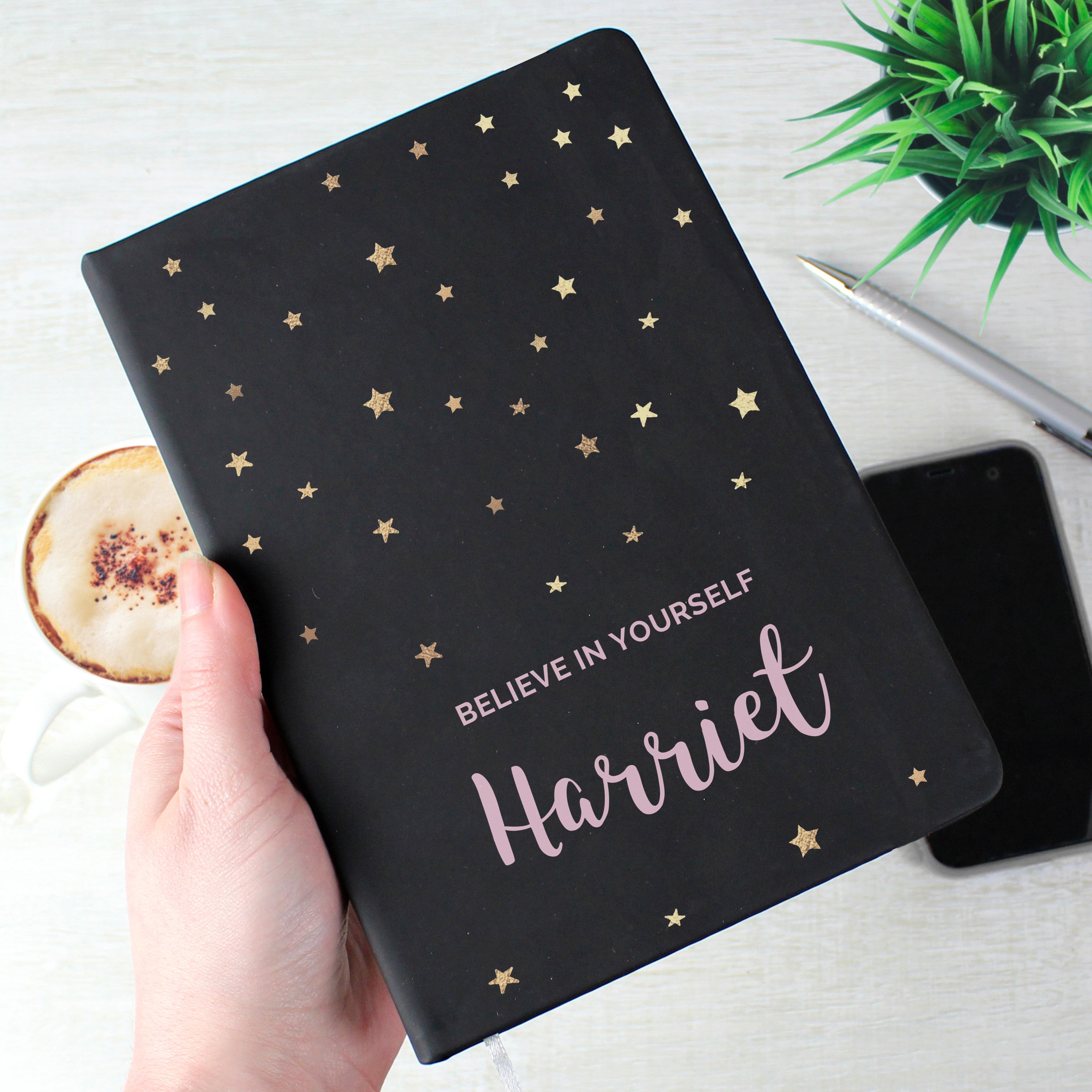 A personalised black hardback notebook with star patterns on the cover, held in a hand, with a mobile phone and a plant in the background.