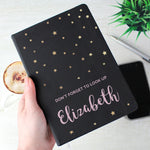 A personalised black hardback notebook with star patterns on the cover, held in a hand, with a mobile phone and a plant in the background.