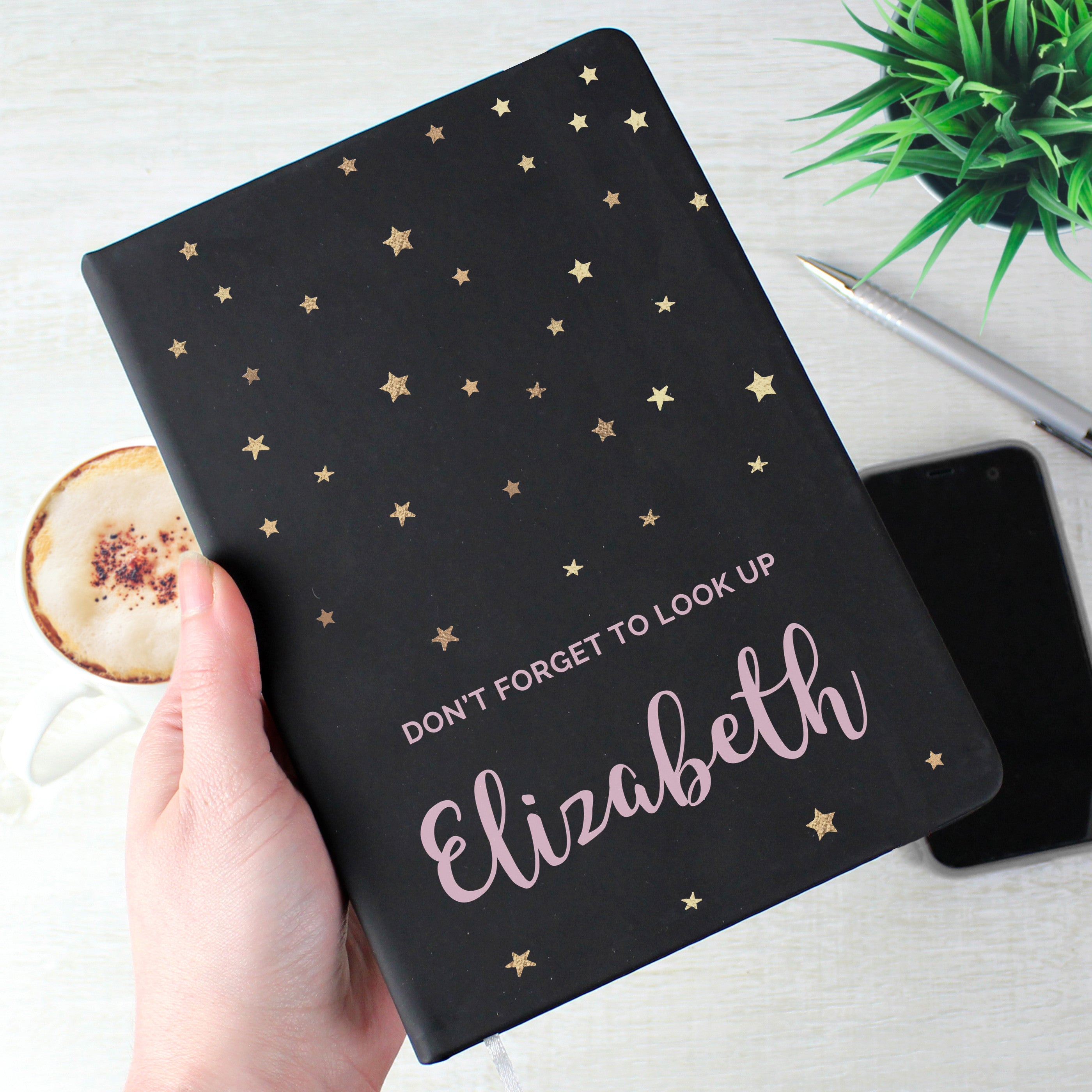 A personalised black hardback notebook with star patterns on the cover, held in a hand, with a mobile phone and a plant in the background.
