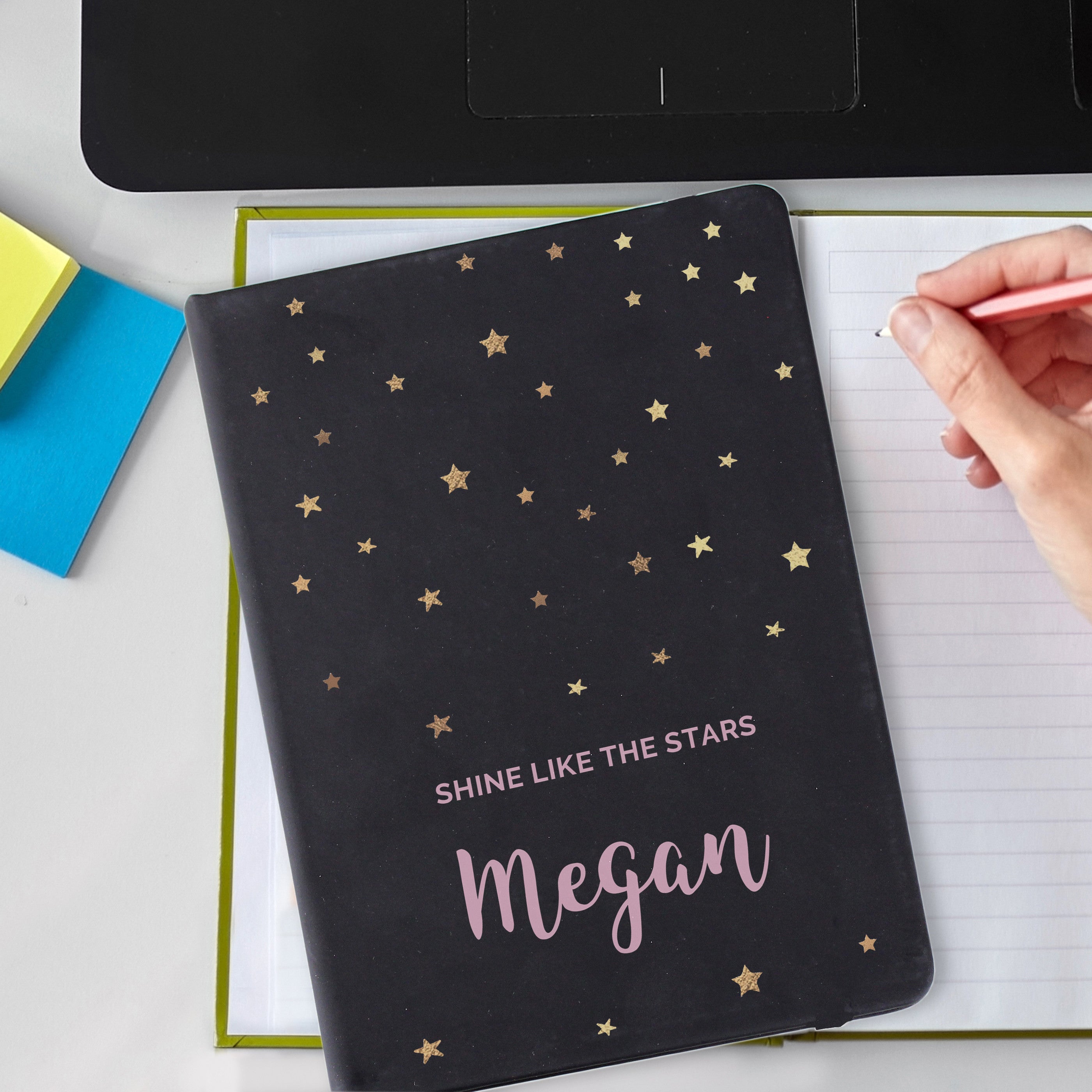 A personalised black hardback notebook with star patterns on the cover, held in a hand, with a mobile phone and a plant in the background.