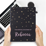 A personalised black hardback notebook with star patterns on the cover, held in a hand, with a mobile phone and a plant in the background.