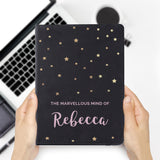 A personalised black hardback notebook with star patterns on the cover, held in a hand, with a mobile phone and a plant in the background.