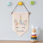 A personalised hanging banner with a unicorn design and the name Samantha printed below it, displayed above a toy rabbit and colorful blocks.