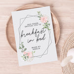 A personalised voucher card with floral design and the text 'This token entitles you to breakfast in bed' along with the name 'Amelia'. The card is displayed on a wooden surface with a round brown plate behind it.