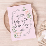 A personalised voucher card with floral design and the text 'This token entitles you to breakfast in bed' along with the name 'Amelia'. The card is displayed on a wooden surface with a round brown plate behind it. 01