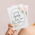 A personalised voucher card with floral design and the text 'This token entitles you to breakfast in bed' along with the name 'Amelia'. The card is displayed on a wooden surface with a round brown plate behind it. 02