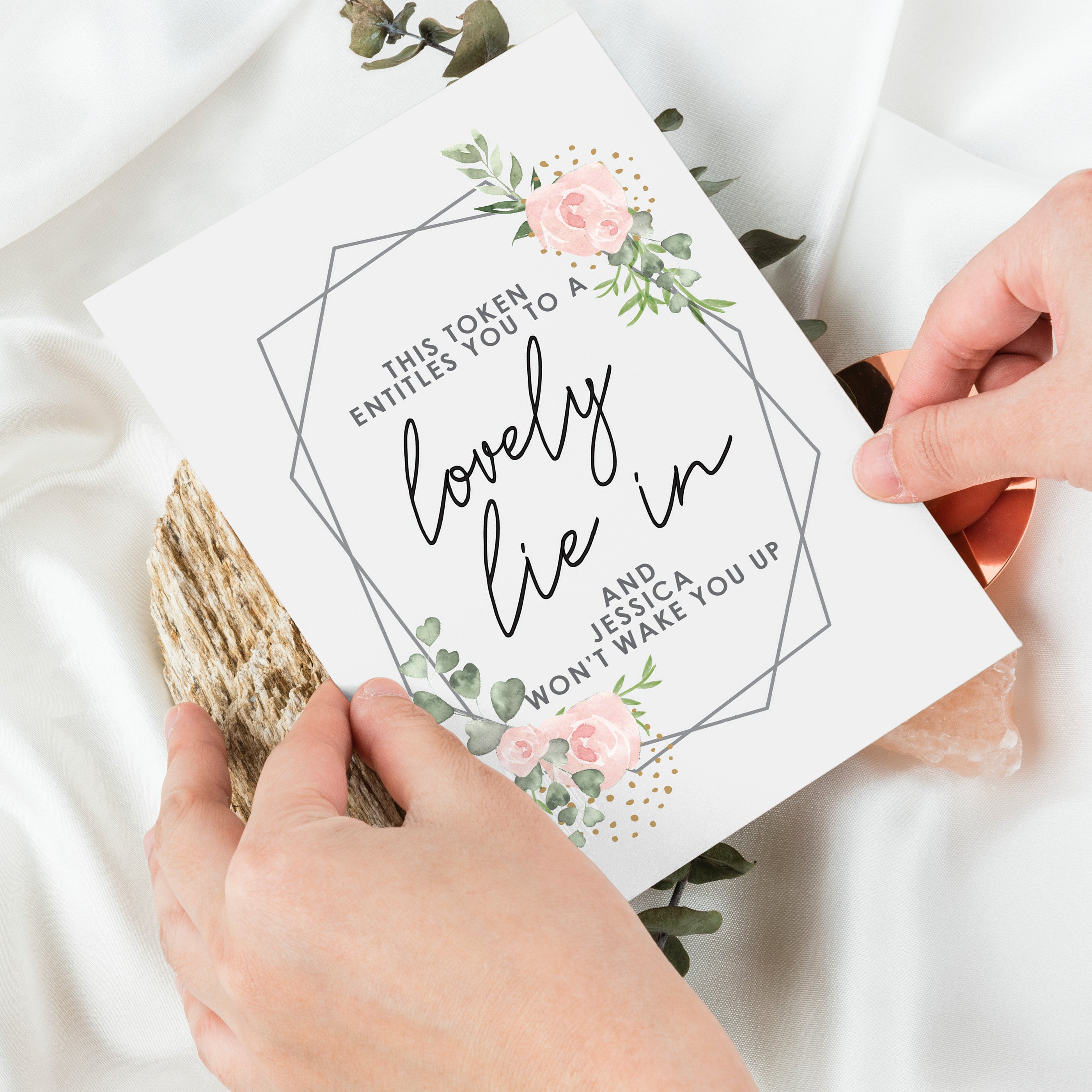 A personalised voucher card with floral design and the text 'This token entitles you to breakfast in bed' along with the name 'Amelia'. The card is displayed on a wooden surface with a round brown plate behind it. 03