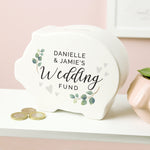 A white ceramic piggy bank with 'Danielle & Jamie's Wedding Fund' printed on the front, adorned with green and brown leaves, placed on a shelf with coins in front.