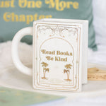 White mug with decorative text and palm trees design, placed on a surface with a blurred book in the background.
