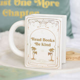 White mug with decorative text and palm trees design, placed on a surface with a blurred book in the background.