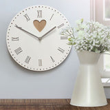 A vintage white wall clock with heart detail displayed on a wall next to a vase with flowers.