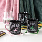 Black mugs with witch-themed text and graphics on a table with a glass and books.
