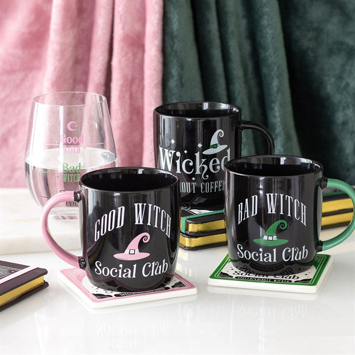 Black mugs with witch-themed text and graphics on a table with a glass and books.