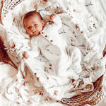 A baby lying on a cream-colored fringe blanket with floral patterns and a visible brand tag on the blanket.