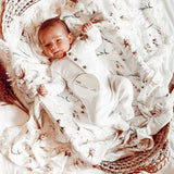 A baby lying on a cream-colored fringe blanket with floral patterns and a visible brand tag on the blanket.
