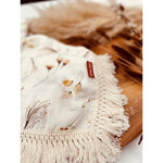 A baby lying on a cream-colored fringe blanket with floral patterns and a visible brand tag on the blanket.