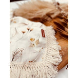 A baby lying on a cream-colored fringe blanket with floral patterns and a visible brand tag on the blanket.
