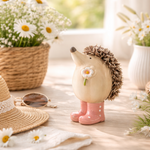 Decorative hedgehog figurine with pink shoes holding a flower, surrounded by flowers and a hat on a wooden surface.