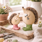 Two decorative hedgehogs with boots on a wooden surface with flowers and eggs in the background.